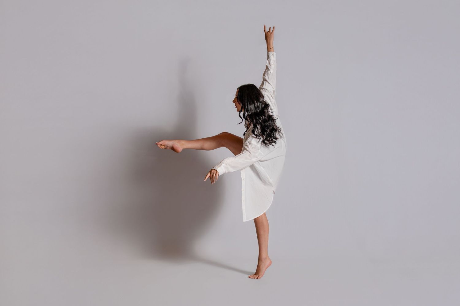 A dancer in a white dress performs an expressive pose with one leg extended back and arm raised upward against a light background.