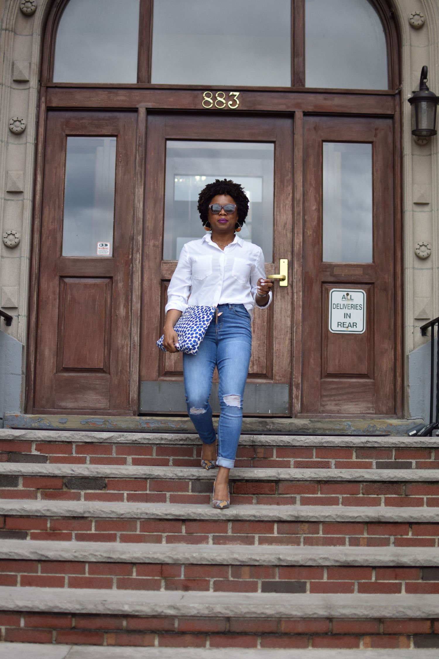 A person sits casually on brick steps outside a brown door in casual attire.