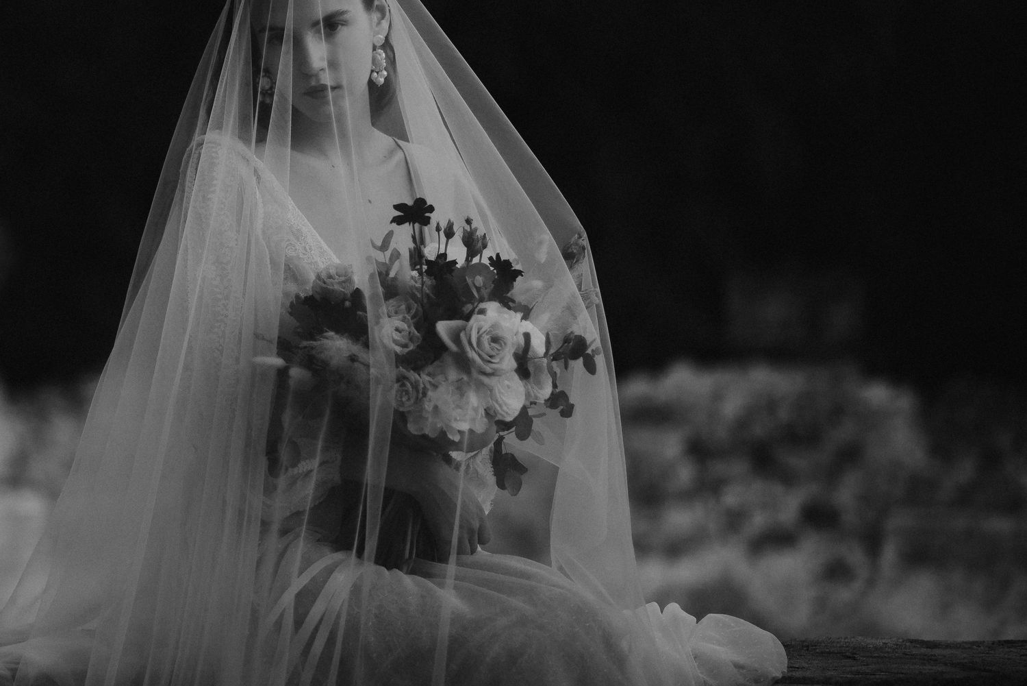 A black and white photograph of a bride in a flowing veil holding a bouquet of flowers in a dreamy outdoor setting.