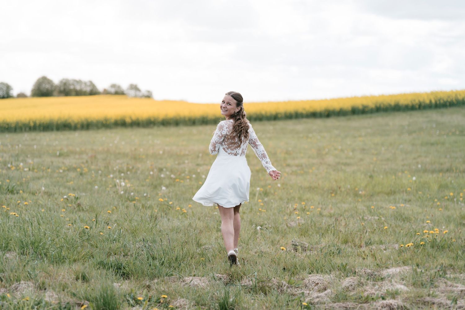 Young woman in white dress twirling in a green field with yellow flowers and trees in the background.