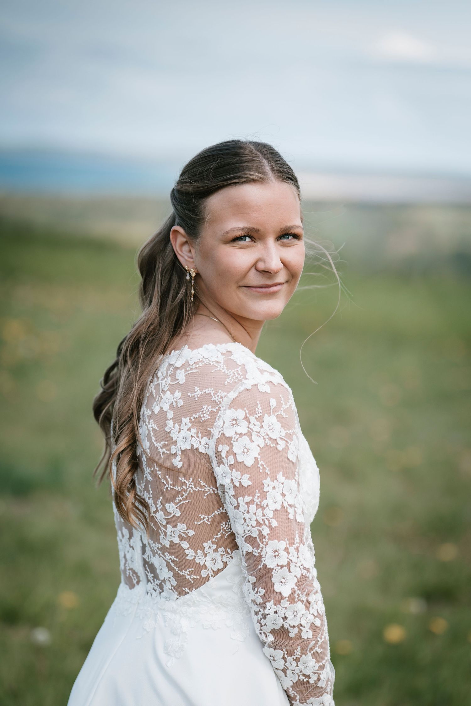 Bride in white lace long-sleeve wedding dress smiling over shoulder outdoors with green hills background.