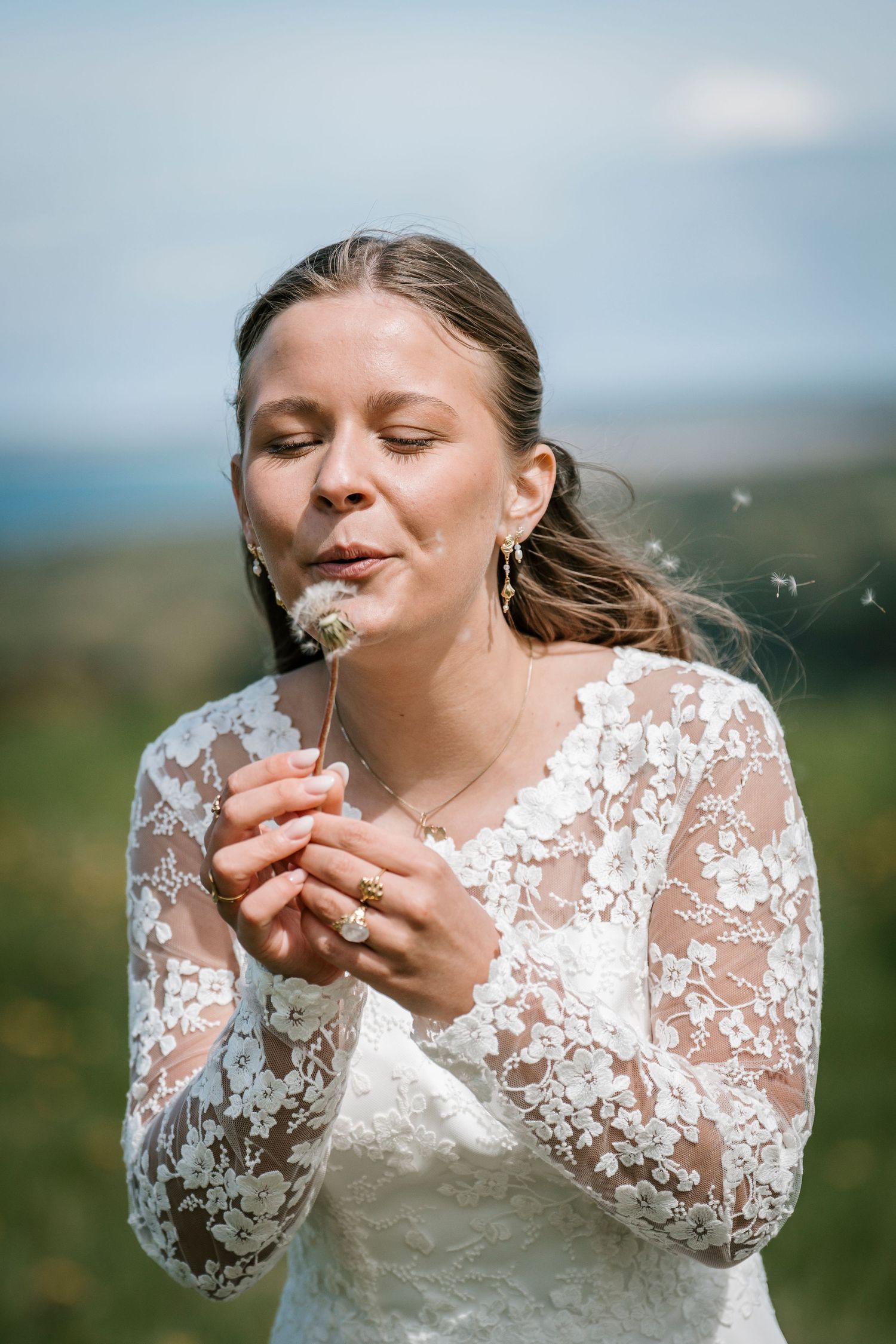 Bride in lace wedding dress blowing dandelion seeds outdoors on a sunny day.
