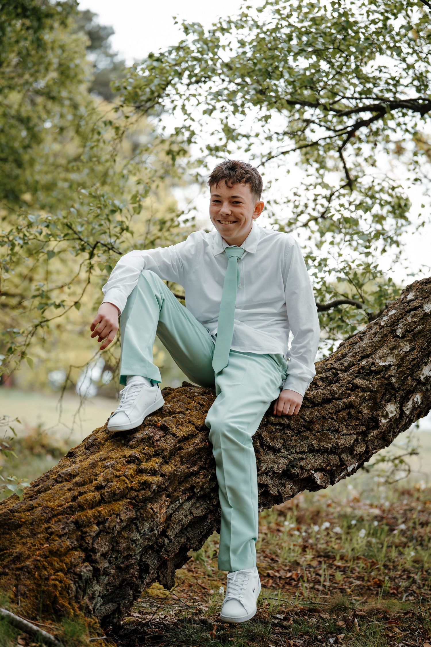 Young man in mint green suit and tie sitting relaxed on a mossy tree branch, smiling outdoors.
