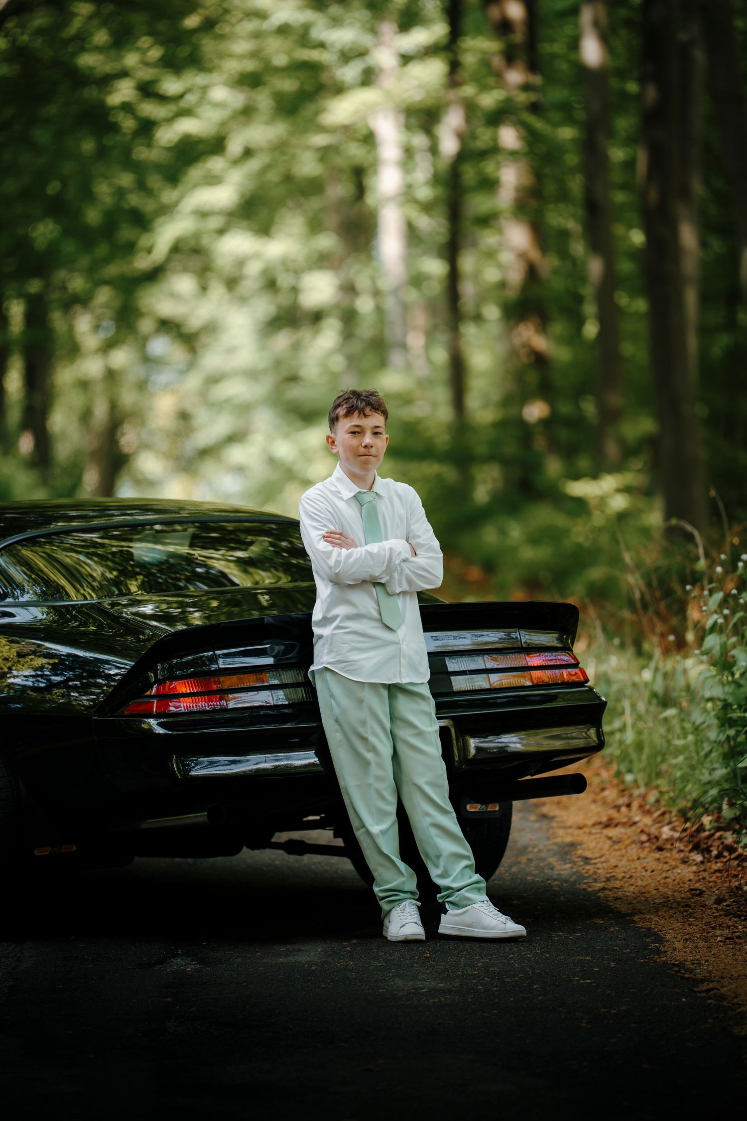 Teen boy in mint green suit leaning against a black classic car on a forest road, arms crossed.