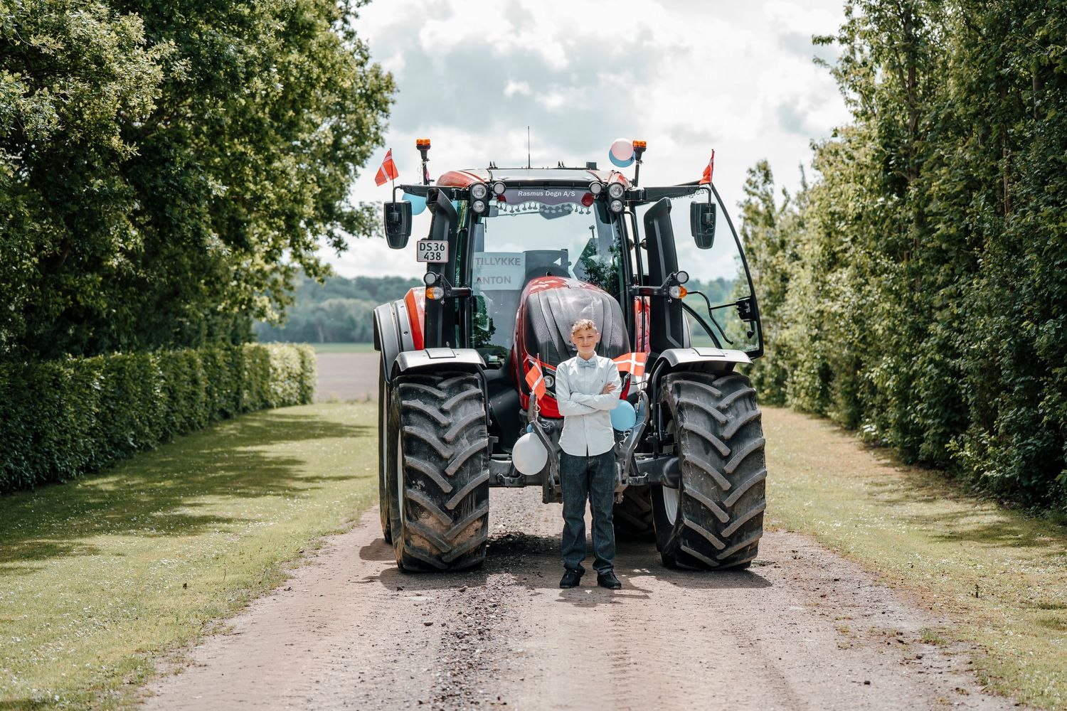 A parent and child admire a large modern red and white tractor on a tree-lined rural path.