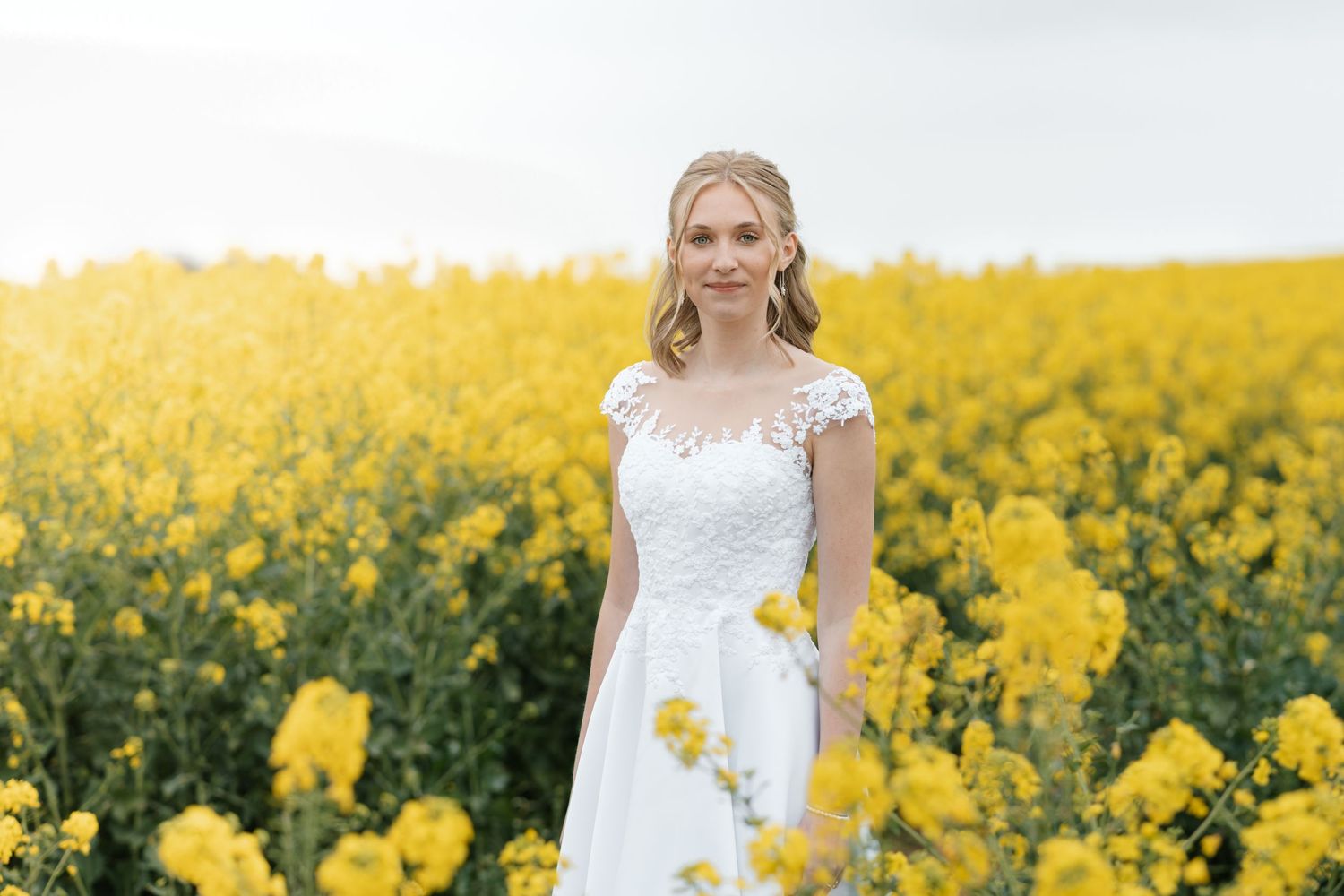 Woman in white lace wedding dress standing in bright yellow canola flower field on overcast day.