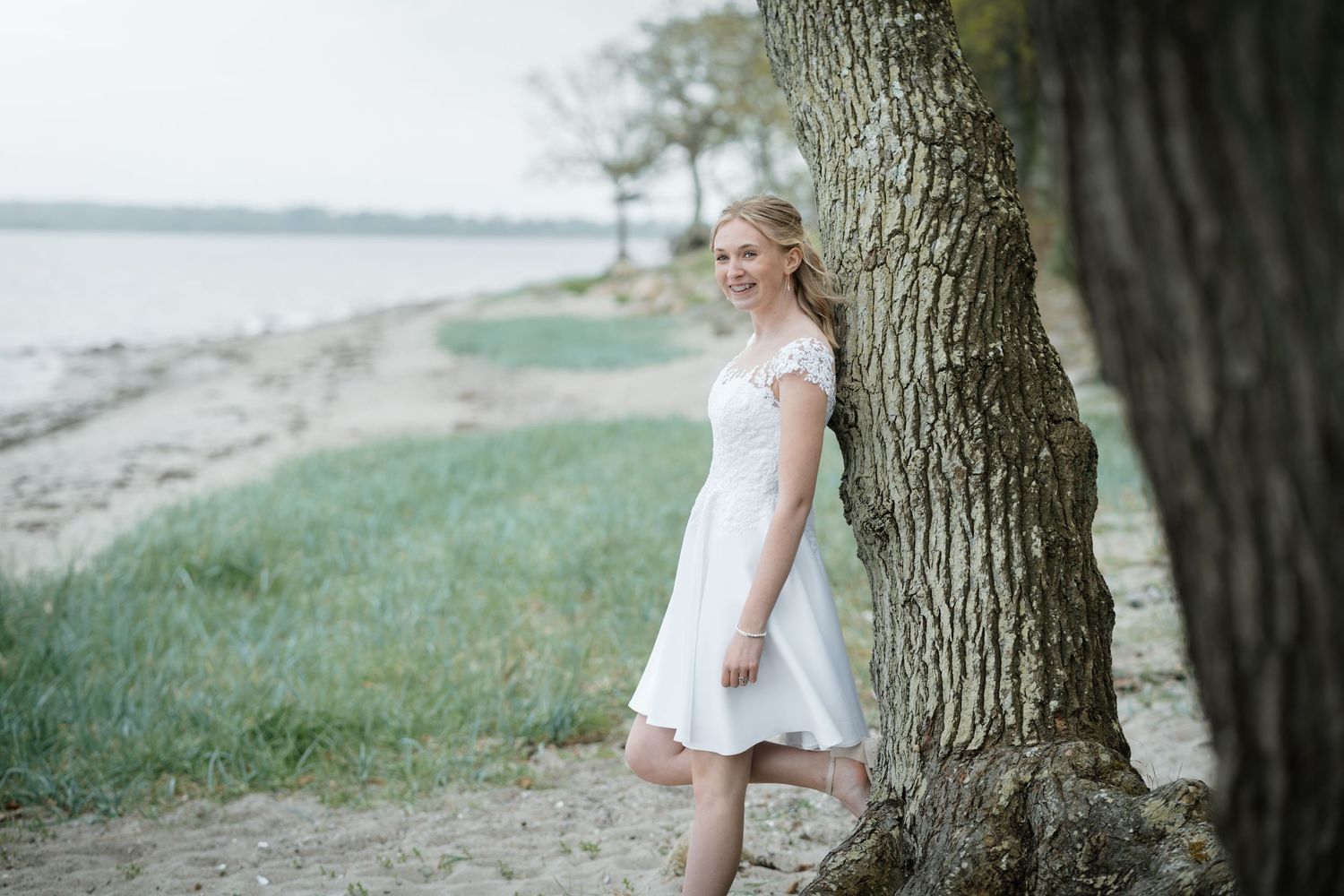 A bride in a white dress leans against a large tree trunk near a serene waterfront on a cloudy day.