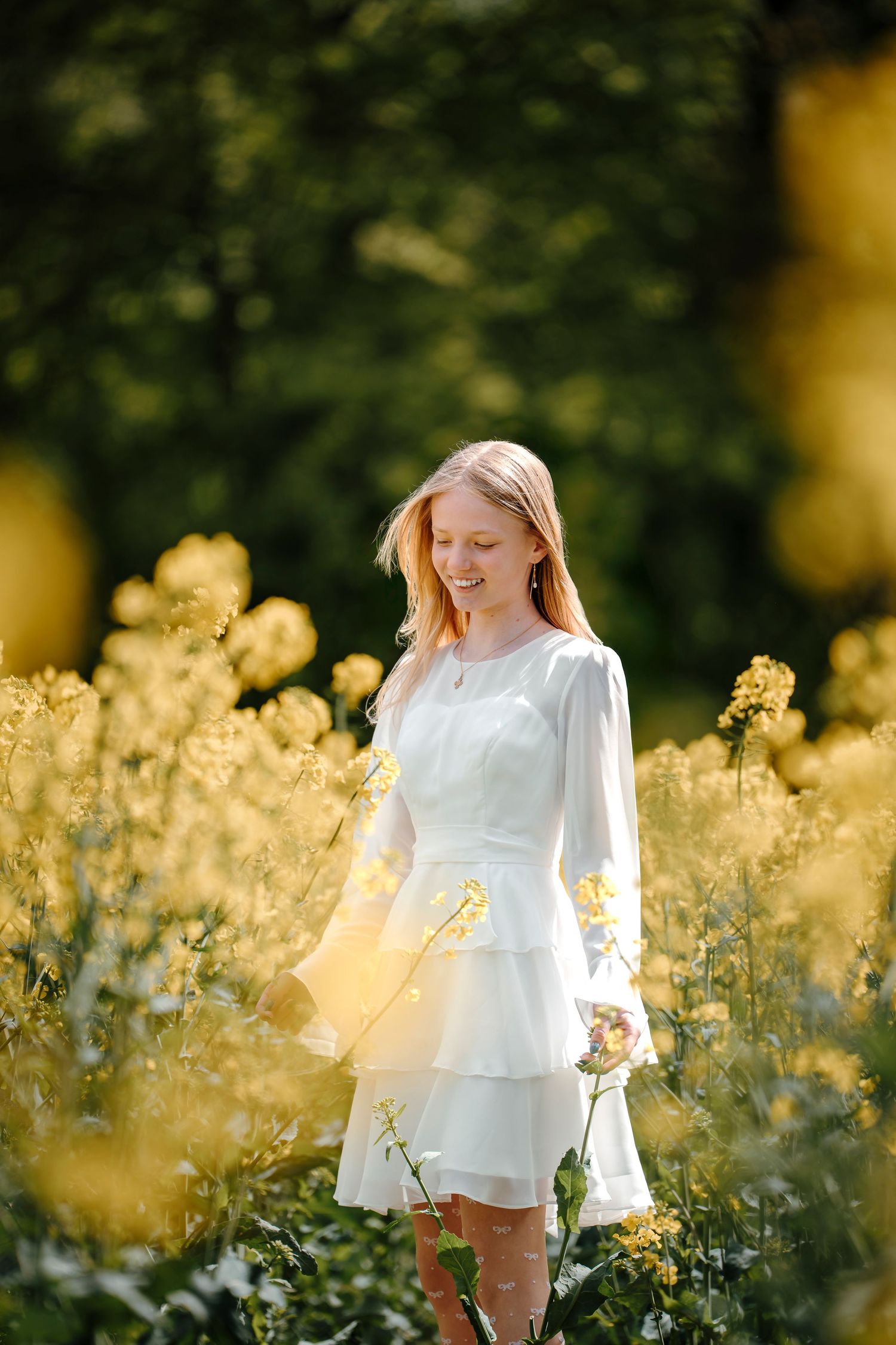 A romantic portrait in a white dress standing among sunny yellow wildflowers in a summer meadow.