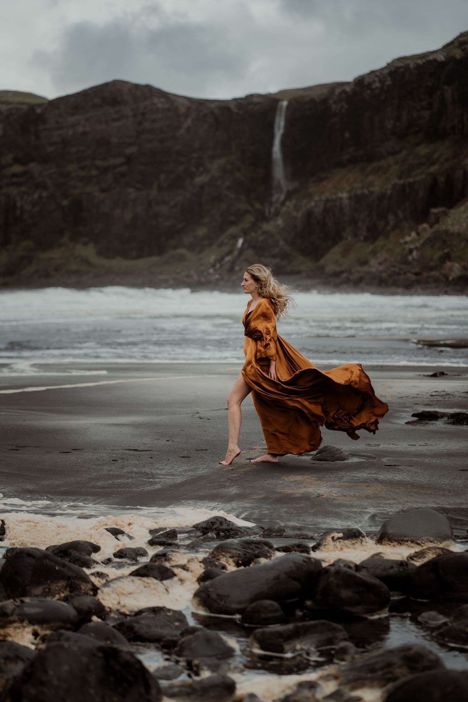 A long flowy rust-colored dress billows in the wind on a dramatic black sand beach with rocky cliffs in the background.