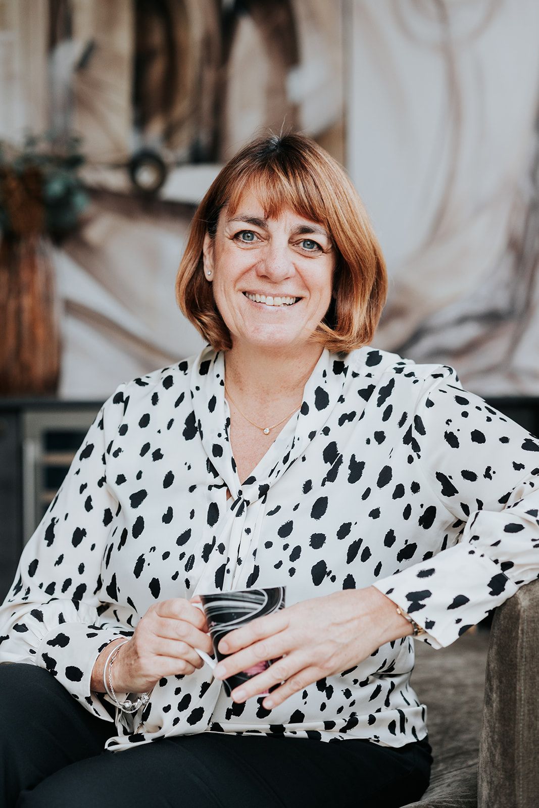 Someone in a black and white polka dot blouse sits casually while holding a drink and smiling at the camera.