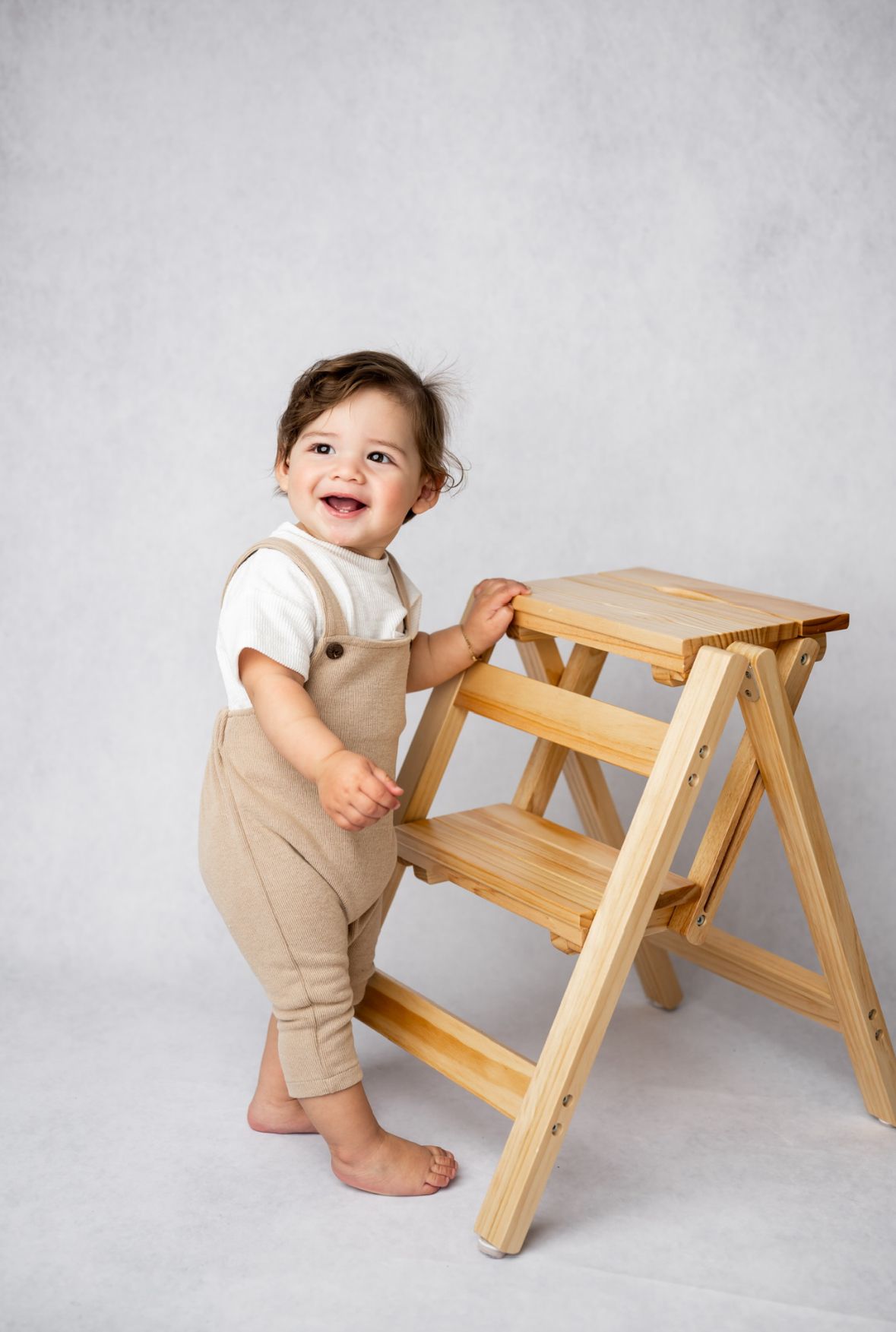 Toddler in beige overalls stands barefoot next to a wooden step stool against a light gray background.