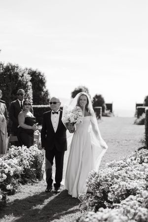 Black and white photograph of bride walking down flower-lined aisle at outdoor ceremony.