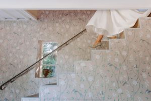White wedding dress flowing down elegant stairs with vintage handrail and floral wallpaper.