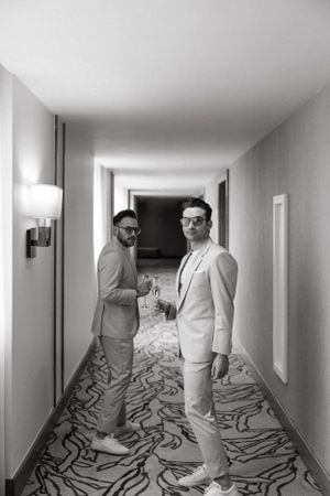 Two men in light-colored suits pose in a modern hotel hallway with patterned carpeting.