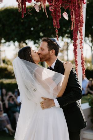 Wedding ceremony kiss under a red floral arch with guests watching.