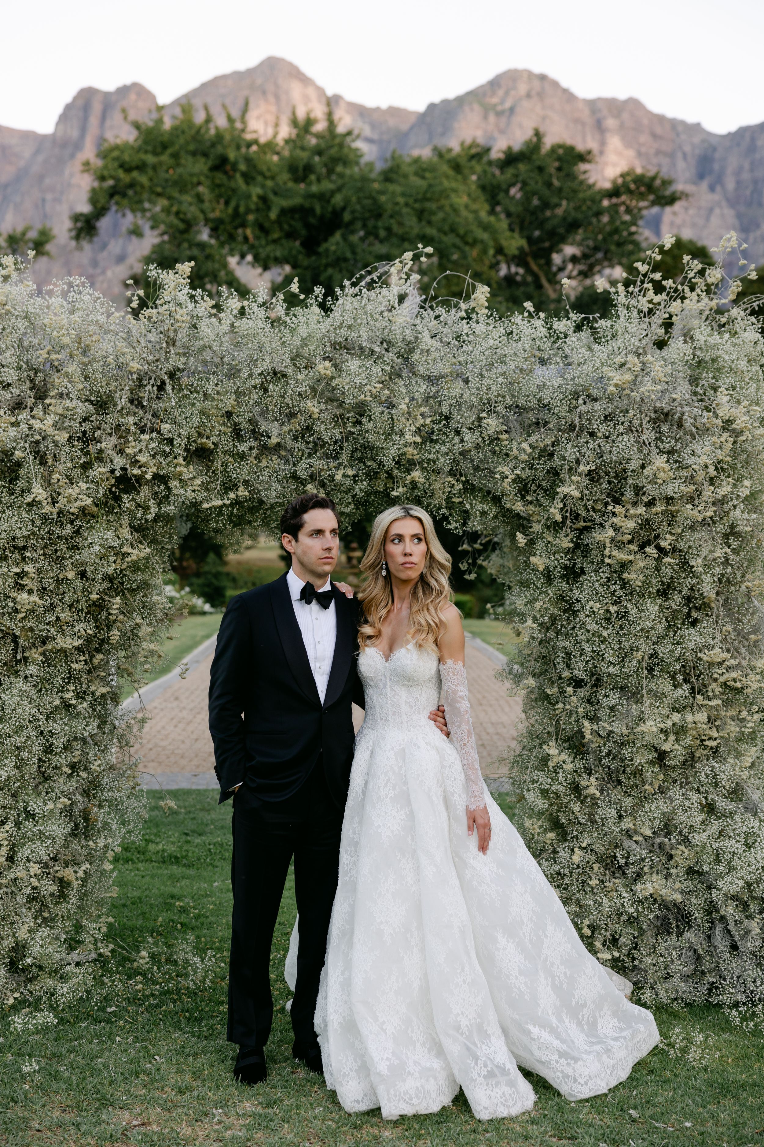 Wedding couple standing in front of blooming white floral arch with mountain backdrop.