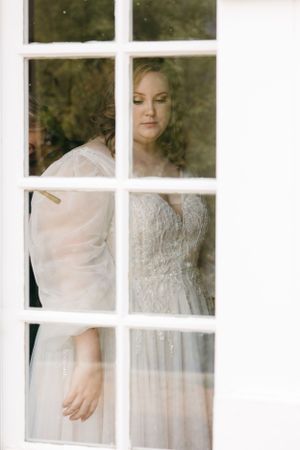 A bride in a lace gown looks through a divided window pane with soft lighting creating a dreamy atmosphere.