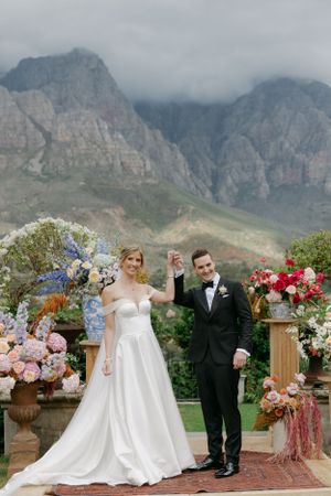 Wedding couple celebrates with raised arms against dramatic mountain backdrop with floral decor.