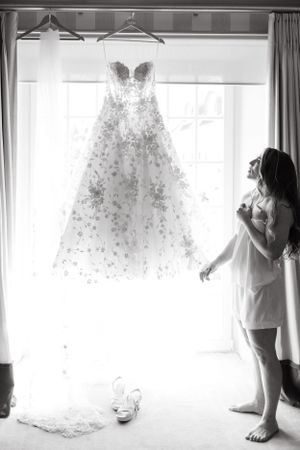 Elegant white wedding dress with floral details hanging by window in morning light.