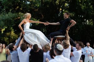 A couple in wedding attire are lifted on shoulders of guests during outdoor celebration at a garden reception.