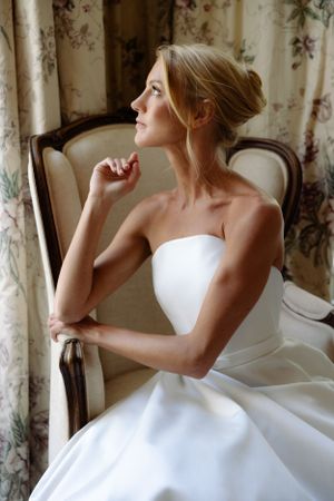 A bride in a classic white strapless gown sits thoughtfully in an elegant chair against floral curtains in soft lighting.