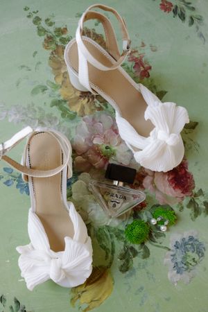 White satin bridal shoes with elegant bow details displayed on floral print background with perfume bottle.