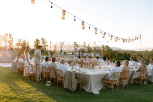 Outdoor wedding reception at sunset with long tables, string lights, and guests dining on a grassy lawn.