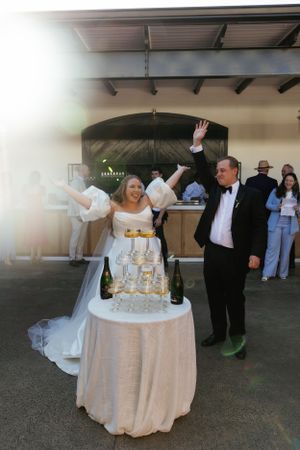 Wedding couple celebrates joyfully with raised arms next to their champagne tower display at outdoor reception.