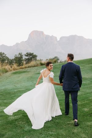 A couple walks on a lush green lawn against a backdrop of majestic mountains at sunset.
