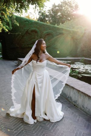 A bride in a stunning white wedding gown with a flowing veil twirls on a garden path during golden hour.