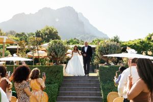 An outdoor wedding ceremony at a scenic venue with mountain backdrop and guests seated on both sides of stone steps.