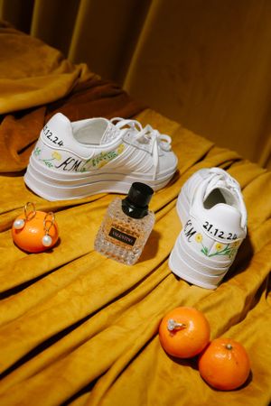 White sneakers with decorative writing displayed on golden fabric alongside oranges and a perfume bottle.