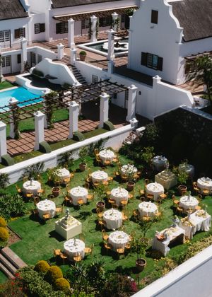 Aerial view of elegant outdoor wedding reception setup with round tables and white umbrellas beside a Cape Dutch style villa with pool.