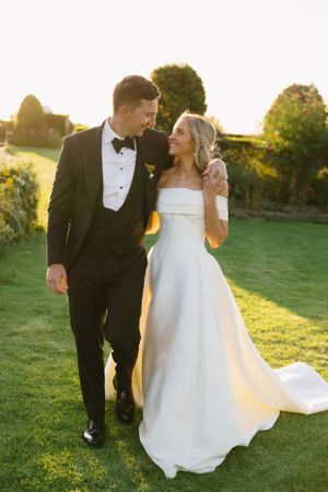 A bride and groom walk together on a lawn at sunset during their wedding, with golden light illuminating the scene.