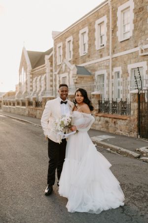 A bride and groom pose together on a street with historic stone buildings in the background.