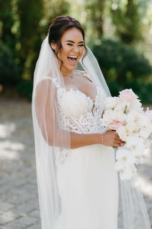 Joyful bride laughing while holding a white bouquet outdoors with sunlit greenery in the background.