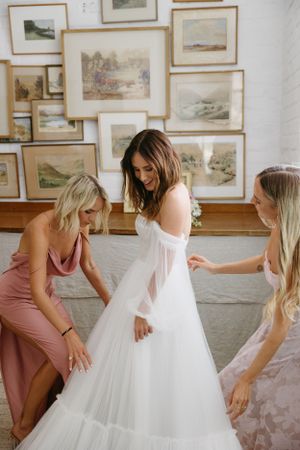 Bridesmaids help adjust a flowing white wedding dress against a wall of framed artwork.