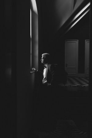 Dramatic black and white photo of a groom looking out window in shadowed interior.