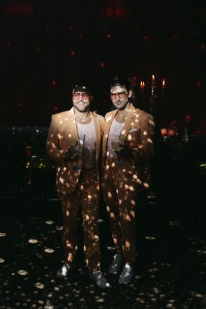 Two people in matching suits stand in a dark room illuminated by sparkling light effects.