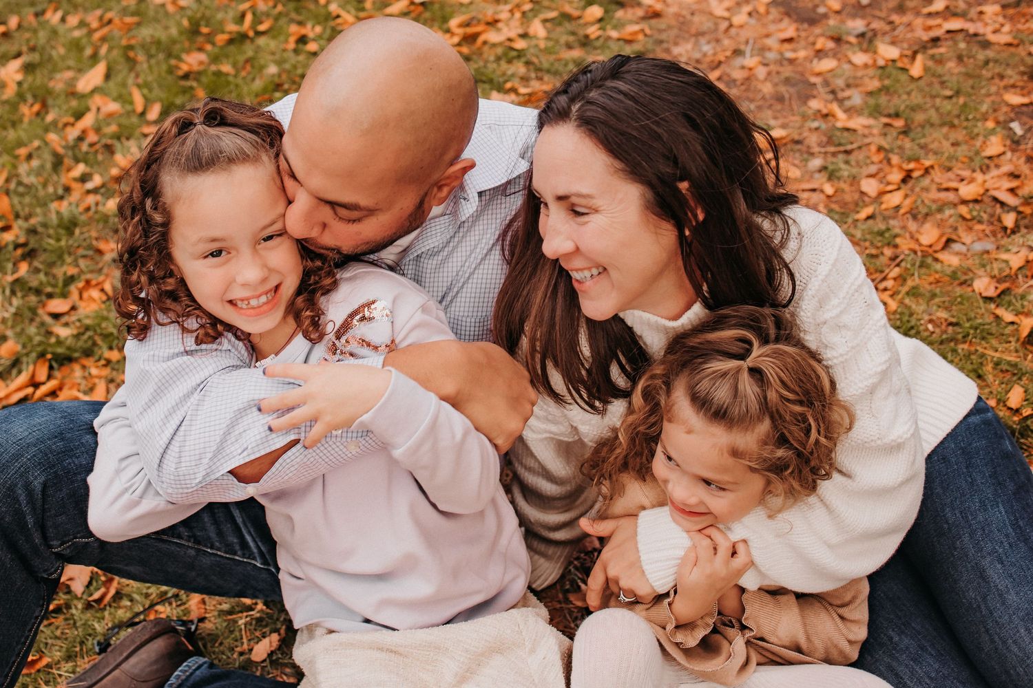 Joyful family laughing together while sitting on autumn leaves in casual clothing during outdoor photoshoot.