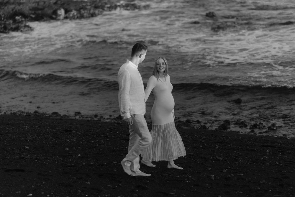 A couple in white outfits stands together on a black sand beach with waves in the background. Maternity photoshoot in Tenerife.