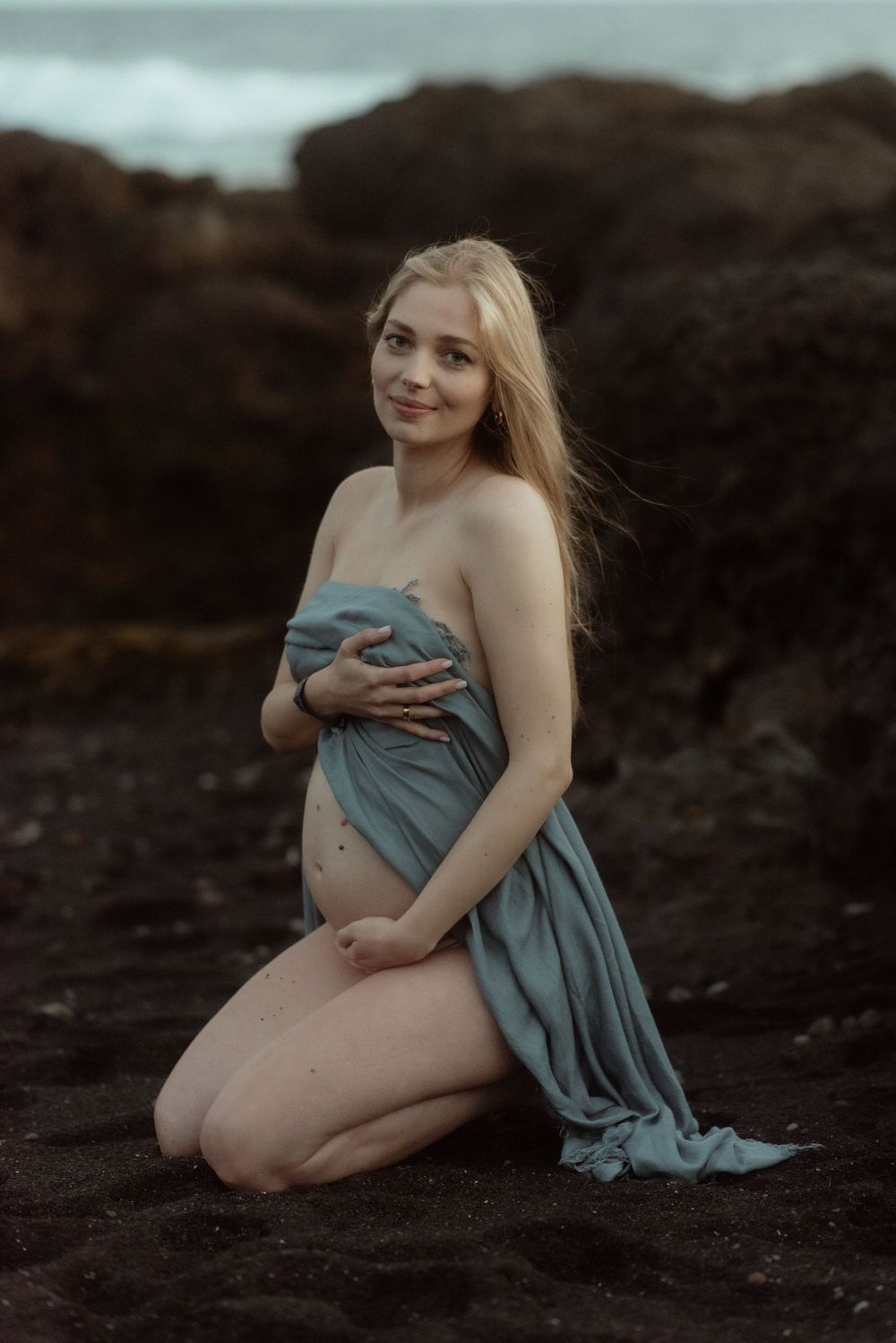 A woman wrapped in blue fabric poses on dark volcanic rocks during a moody outdoor maternity photoshoot in Tenerife.