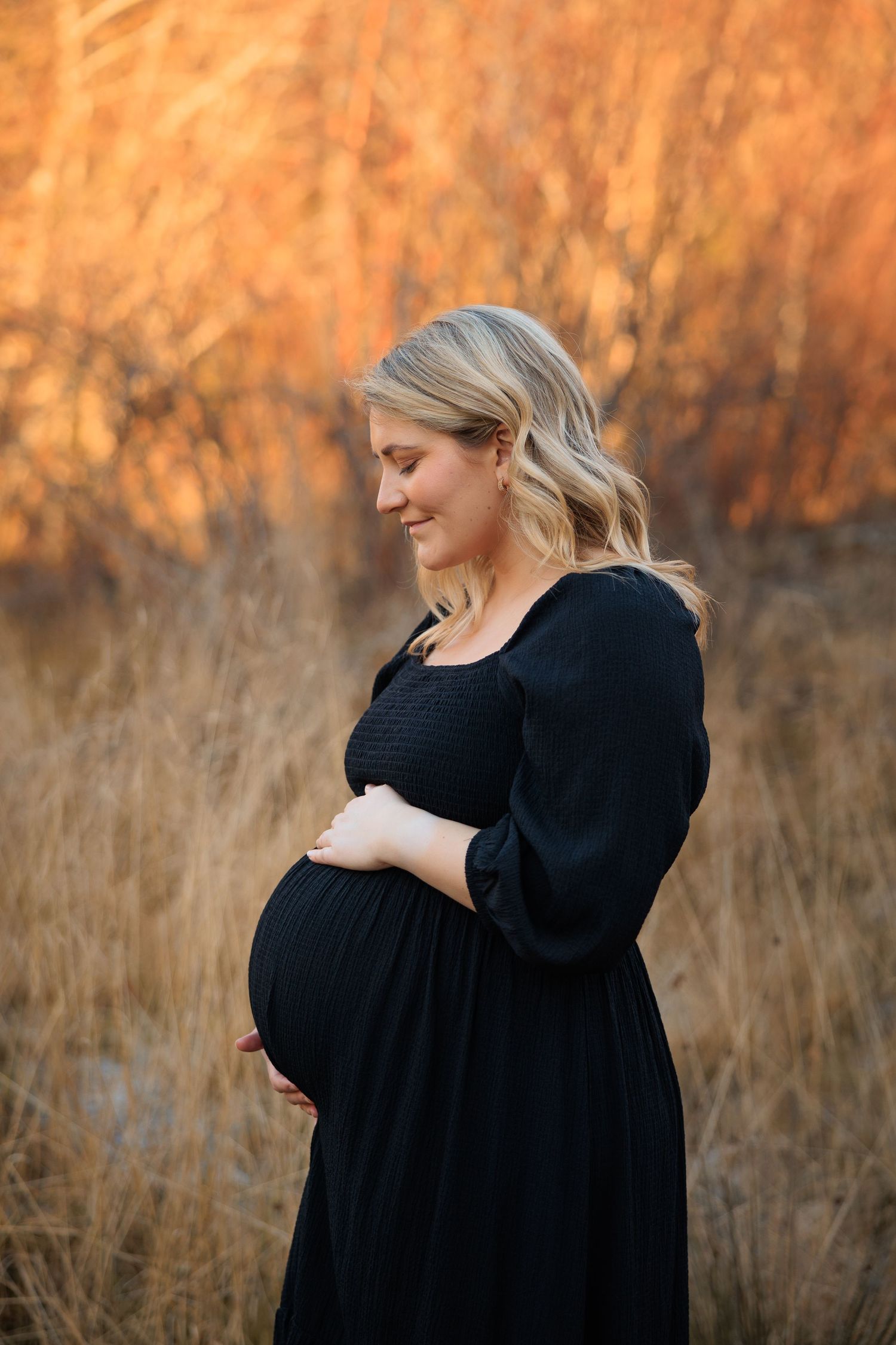 A pregnant woman in a black dress poses in a field during golden hour with warm autumn colors in the background.