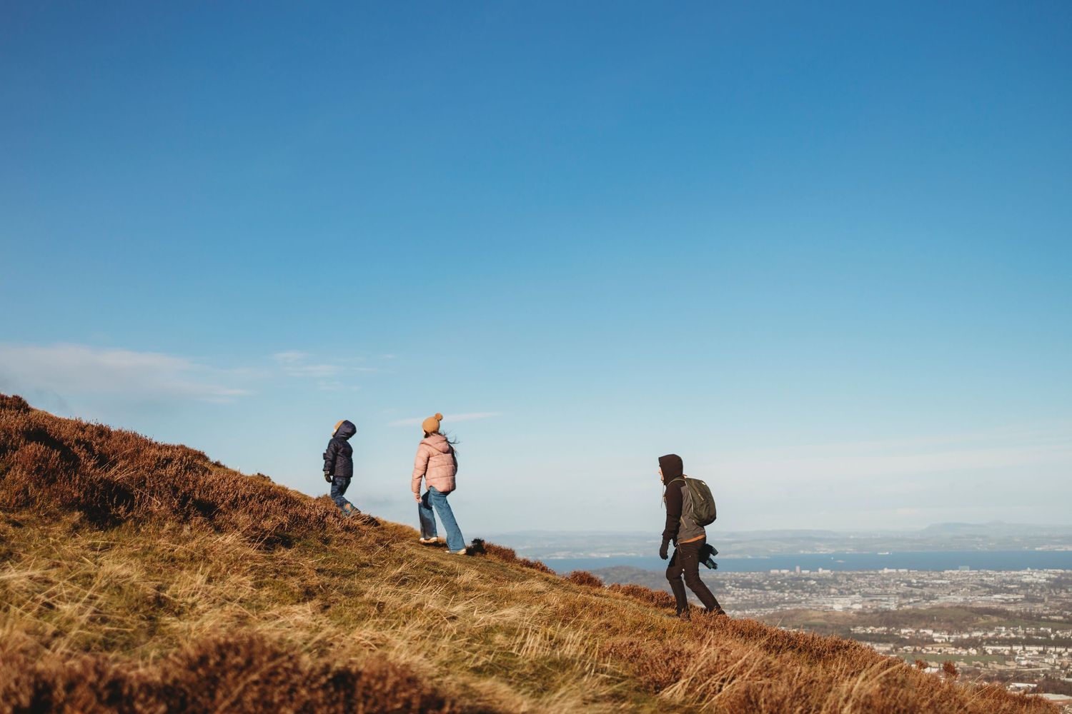 Three hikers walking along a grassy hillside trail overlooking a distant city view on a clear sunny day.