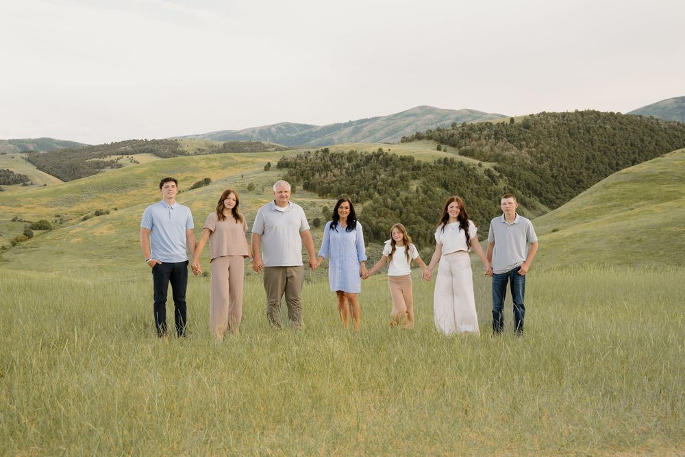 A group of people in light colored clothing stand together in a scenic mountain meadow with rolling green hills.