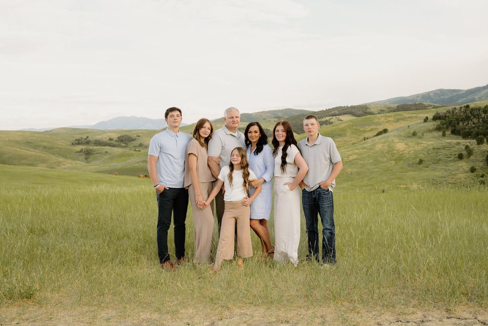 A group of people pose together in a lush green meadow with rolling hills and mountains in the background.