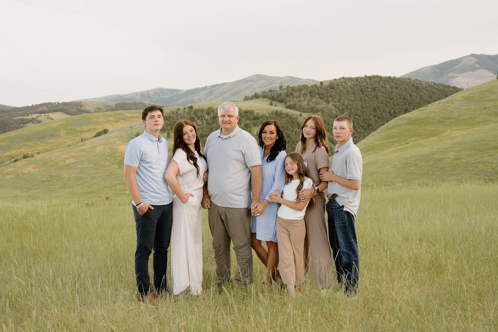 A group poses together in a scenic mountain meadow with rolling green hills and mountains in the background at sunset.