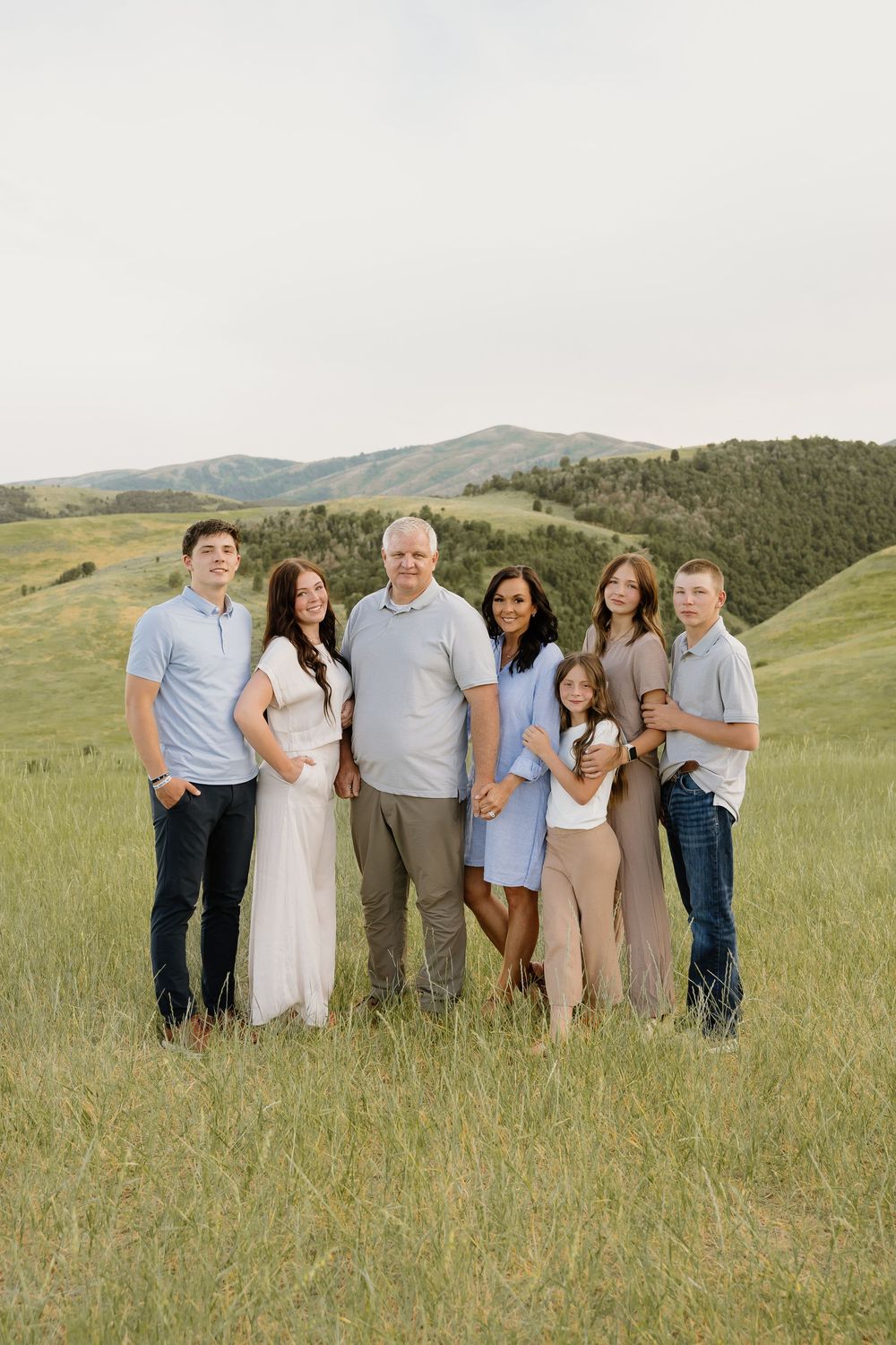 A group of seven people stand together in coordinated blue and neutral outfits against rolling green hills and mountains.