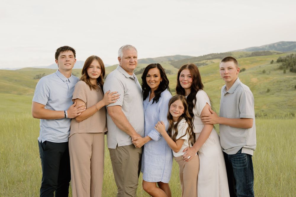 A loving family stands together in casual attire against a scenic backdrop of rolling green hills and mountain landscapes.