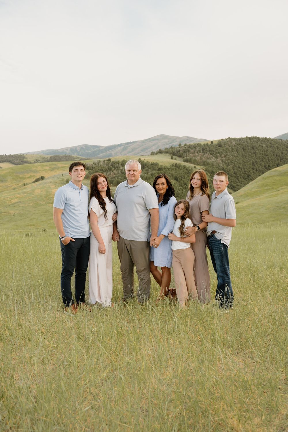 A multi-generational group stands together in a grassy meadow with rolling green hills and mountains in the background.